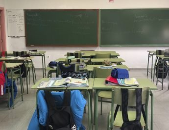 Mesas de un colegio con mochilas en los asientos y una pizarra al final.