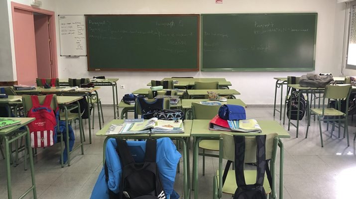 Mesas de un colegio con mochilas en los asientos y una pizarra al final.