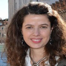 Primer plano de Lola Pons sonriendo con el pelo rizado. La mujer lleva un collar anudado y unos pendientes largos, además de una camisa blanca y una chaqueta verde oscuro. El fondo de la imagen es un conjunto de edificios, uno más alto blanco y dos más bajos de color marrón.