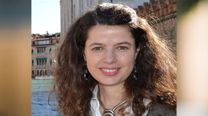 Primer plano de Lola Pons sonriendo con el pelo rizado. La mujer lleva un collar anudado y unos pendientes largos, además de una camisa blanca y una chaqueta verde oscuro. El fondo de la imagen es un conjunto de edificios, uno más alto blanco y dos más bajos de color marrón.