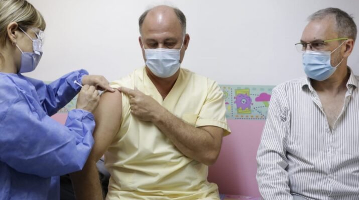 En la imagen aparecen tres personas. Una mujer y dos hombres, todos con mascarilla. La mujer es la sanitaria que se está encargando de administrarle la vacuna a uno de los hombres, el otro permanece al lado de ellos mirándoles. La mujer viste con un traje de seguridad color azul, uno de los hombres con una camisa y pantalones color amarillo claro y el otro hombre lleva una camisa de rayas. Al fondo, la pared es rosa claro y blanca con una especie de dibujos animados pintados en ella.