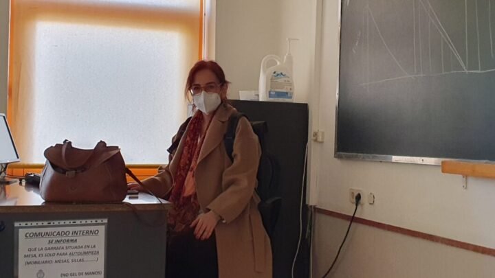 María Nieves San Emeterio posa sentada en la mesa del profesor, con mascarilla. Detrás de ella se encuentra la pizarra en la que ha dibujado gráficas.