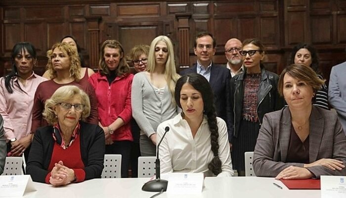 Manuela Carmena, ex-alcaldesa de Madrid sentada en una mesa junto a dos mujeres trnas. Detrás de ellas, más personas del colectivo. Todas ellas detrás de los micros de la rueda de prensa.