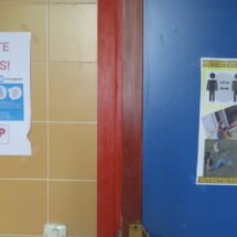 2 carteles, uno en un apuerta azul y otra en una pared de azulejo naranja que muestran medidas para no contagiar