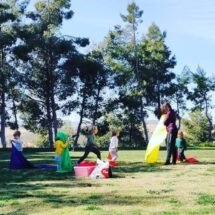 Cinco niños realizando aprendizaje experimental en un prado rodeado de árboles junto a su mamá de día