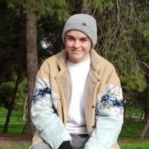 Pablo sonriente vestido con un gorro gris y una chaqueta de color marrón y azul cielo. De fondo se ve un paisaje con árboles.