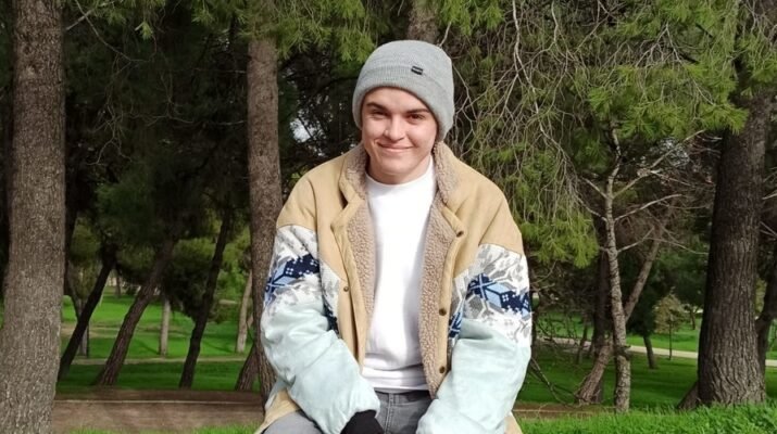 Pablo sonriente vestido con un gorro gris y una chaqueta de color marrón y azul cielo. De fondo se ve un paisaje con árboles.