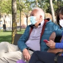 Plano medio de una pareja adulta de un hombre y una mujer sentados en el banco de un parque. Ambos llevan mascarilla.