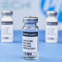 Tres frascos del compuesto de Pfizer y BioNTech contra la COVID19 sobre una superficie lisa azul. En el fondo, blanco y difuminado, se intuye el logotipo de la farmacéutica de color azul.