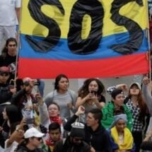 Plano picado y frontal de un grupo de unas veinte personas colombianas manifestándose con una gran bandera de Colombia en la que aparece escrito en letras mayúsculas SOS.