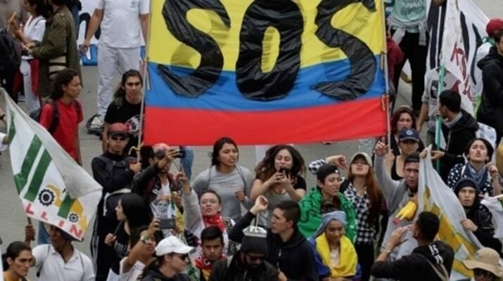 Plano picado y frontal de un grupo de unas veinte personas colombianas manifestÃ¡ndose con una gran bandera de Colombia en la que aparece escrito en letras mayÃºsculas SOS.