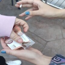 En la imagen aparecen las manos de dos personas en un negocio de compraventa de droga propia del mercado negro. Una de las manos sostiene un billete de 10 euros y una bolsita de droga. La otra mano está cogiendo uno de los dos elementos.