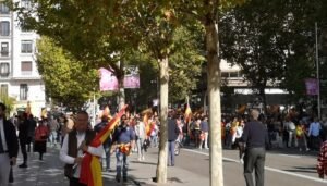 Personas caminando hacía la manifestación de Vox en los alrededores de la Plaza de Colón. En la parte superior se ven las hojas de los árboles, las personas que caminan llevan banderas de España.