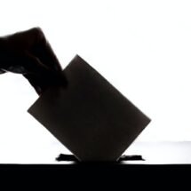 Foto en blanco y negro. Una mano introduce el sobre de su voto en una urna electoral.