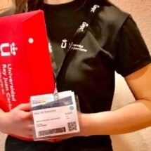 Una persona con merchandising de la Universidad Rey Juan Carlos para ejercer de orientadora en la Feria AULA. Lleva una camiseta negra de manga corta, una acreditación blanca con un código QR y porta en sus manos una carpeta roja.