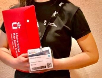 Una persona con merchandising de la Universidad Rey Juan Carlos para ejercer de orientadora en la Feria AULA. Lleva una camiseta negra de manga corta, una acreditación blanca con un código QR y porta en sus manos una carpeta roja.