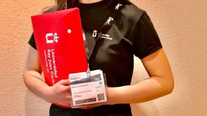 Una persona con merchandising de la Universidad Rey Juan Carlos para ejercer de orientadora en la Feria AULA. Lleva una camiseta negra de manga corta, una acreditación blanca con un código QR y porta en sus manos una carpeta roja.