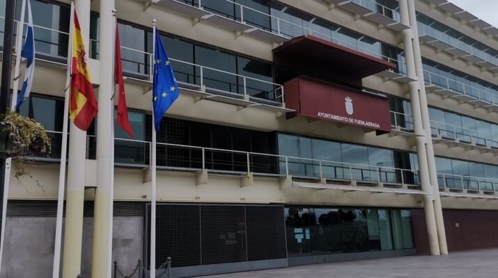 Fachada del ayuntamiento de Fuenlabrada. Se ven tres banderas. El edificio cuenta con numerosos balcones y ventanas acristaladas. En uno de ellos se ve el escudo de Fuenlabrada.