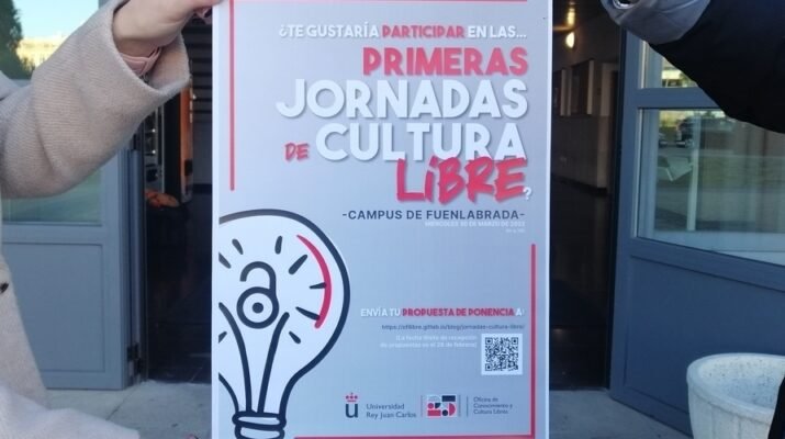 Cartel promocional de las Primeras Jornadas de Cultura Libre de la Universidad Rey Juan Carlos, sostenido por dos personas diferentes a las que solo se les ven los brazos frente a las puertas abiertas del edificio de Gestión del campus, donde se celebrará el evento