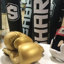 Espejos en la parte de atrás en el gimnasio de boxeo que refleja los sacos de boxeo de color negro con letras blancas, una mesa blanca y encima un guante de boxeo de color dorado y un casco rojo y blanco