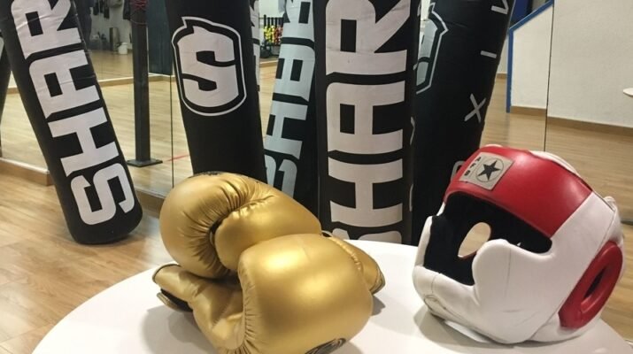 Espejos en la parte de atrás en el gimnasio de boxeo que refleja los sacos de boxeo de color negro con letras blancas, una mesa blanca y encima un guante de boxeo de color dorado y un casco rojo y blanco