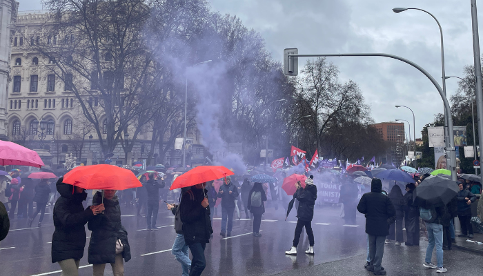 Plano general de humo violeta esparcido en la manifestación del 8M de Madrid con los manifestantes y varios transeúntes.