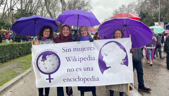 Cuatro mujeres sonrientes sostienen una pancarta que dice "Sin mujeres, Wikipedia no es una enciclopedia" durante la manifestación feminista.