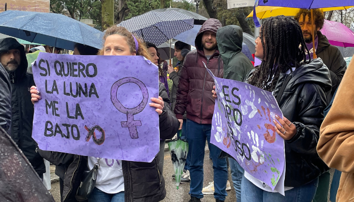 Cartel morado mujer "Si quiero la luna me la bajo yo". Paraguas y gente alrededor.