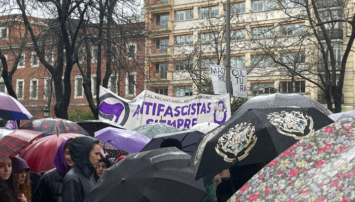 Pancarta blanca y morada al fondo con leyenda "antifascistas siempre", alrededor de paraguas y gente secundando la manifestación.