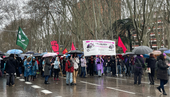 Plano general de manifestantes con pancarta grande "Me cuidan mis amigas y no la policía". Carteles de símbolos y banderas de partidos políticos y organizaciones.