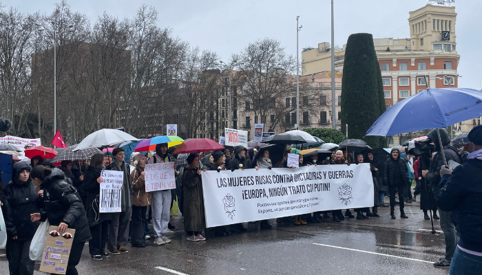 Manifestantes por mujeres rusas, con paraguas y pancarta grande horizontal "Las mujeres rusas contra dictaduras y guerras ¡Europa, ningún trato con Putin!". Personas manifestantes con carteles frente a un cámara que graba el mensaje de su marcha.