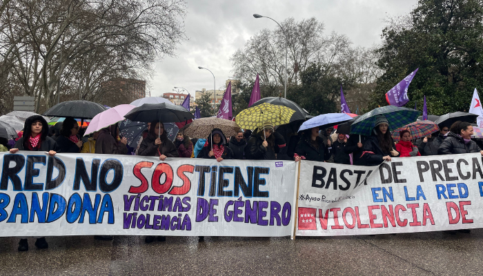 Personas con paraguas sostienen pancartas que denuncian la falta de apoyo a víctimas de violencia de género en una manifestación feminista.