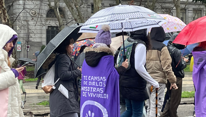 Mujer con capa morada que dice "¿Te cansas de oírlo?, ¡nosotras de vivirlo!", acompañada de personas con paraguas en una manifestación feminista.