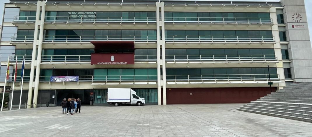 Plano general de la fachada del ayuntamiento de Fuenlabrada. Donde resalta el balcón de actos y un piso mas abajo a la izquierda esta colgado una pancarta del 8m; un grupo de mujeres a la izquierda al fondo; y más al fondo una camioneta en la entrada del edificio la cual es blanca.