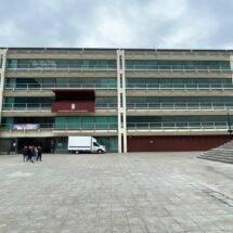 Plano general de la fachada del ayuntamiento de Fuenlabrada. Donde resalta el balcón de actos y un piso mas abajo a la izquierda esta colgado una pancarta del 8m; un grupo de mujeres a la izquierda al fondo; y más al fondo una camioneta en la entrada del edificio la cual es blanca.