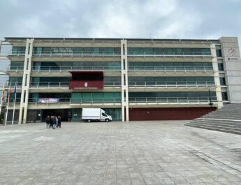 Plano general de la fachada del ayuntamiento de Fuenlabrada. Donde resalta el balcón de actos y un piso mas abajo a la izquierda esta colgado una pancarta del 8m; un grupo de mujeres a la izquierda al fondo; y más al fondo una camioneta en la entrada del edificio la cual es blanca.