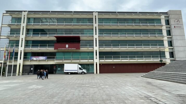 Plano general de la fachada del ayuntamiento de Fuenlabrada. Donde resalta el balcón de actos y un piso mas abajo a la izquierda esta colgado una pancarta del 8m; un grupo de mujeres a la izquierda al fondo; y más al fondo una camioneta en la entrada del edificio la cual es blanca.
