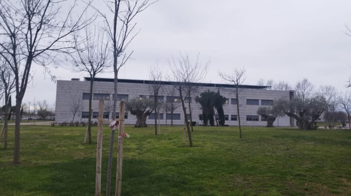 La imagen muestra un plano opanorámico del campus de Feunlabrada. Hay césped colorido y alho húmedo, un edificio grisáceo y rectángular frente el objetivo y varios árboles esbeltos a lo largo del plano. Asimismo el cielo está completamente encapotado.