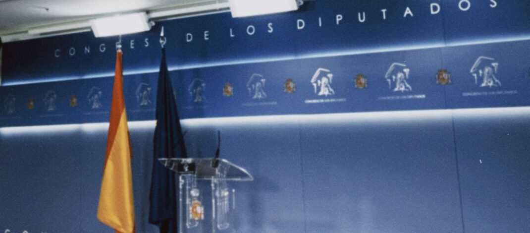 Sala de prensa del congreso de los diputados dónde se ve el fondo azul, la bandera de España y de la UE y un atril transparente, solo muestra la parte de la ponencia, no la parte de prensa