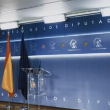 Sala de prensa del congreso de los diputados dónde se ve el fondo azul, la bandera de España y de la UE y un atril transparente, solo muestra la parte de la ponencia, no la parte de prensa
