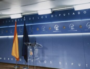 Sala de prensa del congreso de los diputados dónde se ve el fondo azul, la bandera de España y de la UE y un atril transparente, solo muestra la parte de la ponencia, no la parte de prensa