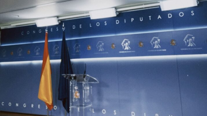 Sala de prensa del congreso de los diputados dónde se ve el fondo azul, la bandera de España y de la UE y un atril transparente, solo muestra la parte de la ponencia, no la parte de prensa