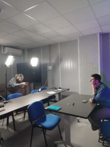 Imagen d de la sala Lady Gaga en la Casa de la música de Fuenlabrada que sirve como estudio para grabar el podcast No todo lo vale con, Óscar Blanco, uno de los presentadores del programa sentado en la mesa del decorado y la entrevistadora fuera de cámara fotografiando al presentador.