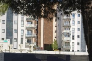 Edificios de varias plantas, hay un árbol a la derecha que tapa parte del edificio y la parte superior de la imagen con sus hojas. En la parte de abajo de la imagen se puede ver una valla de obras con el nombre de una empresa recortada, mientras que en algunos balcones del edificio que mencionaba anteriormente, se puede leer "No a la gentrificación"