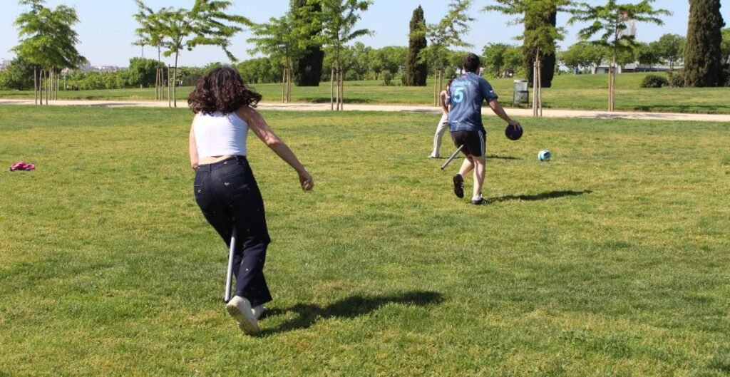 Hay tres personas en la fotografía, todas están con el palo de PVC que simula la escoba. Hay una chica corriendo para atrapar atrapar la quaffle, mientras que un chicho intenta darle a otro con la bludger.