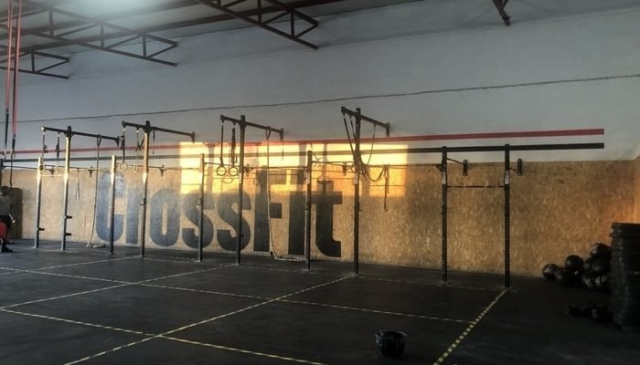 Un gimnasio de CrossFit vacío y en penumbra. No hay espejos porque el CrossFit no es un deporte que se quiera fijar en la estética. Aparece el logo de CrossFit pintado de negro en la pared, que es de caucho.