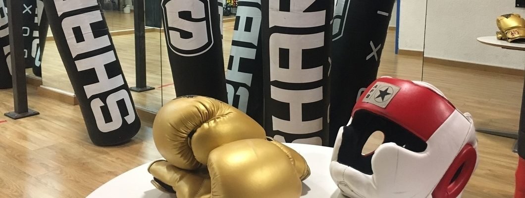 Espejos en la parte de atrás en el gimnasio de boxeo que refleja los sacos de boxeo de color negro con letras blancas, una mesa blanca y encima un guante de boxeo de color dorado y un casco rojo y blanco