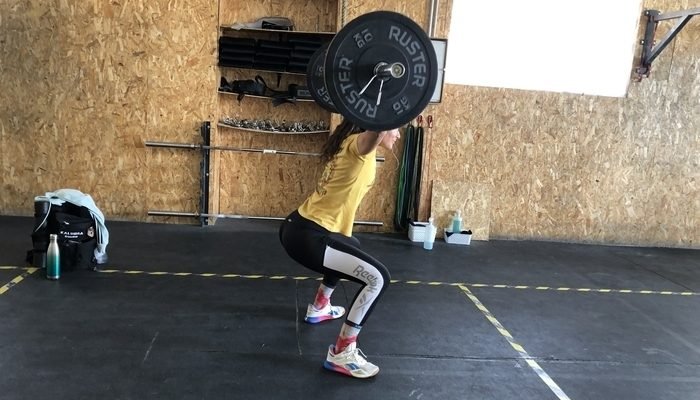 Aparece una chica joven con camiseta amarilla y leggins negros levantando unas pesas al practicar CrossFit.